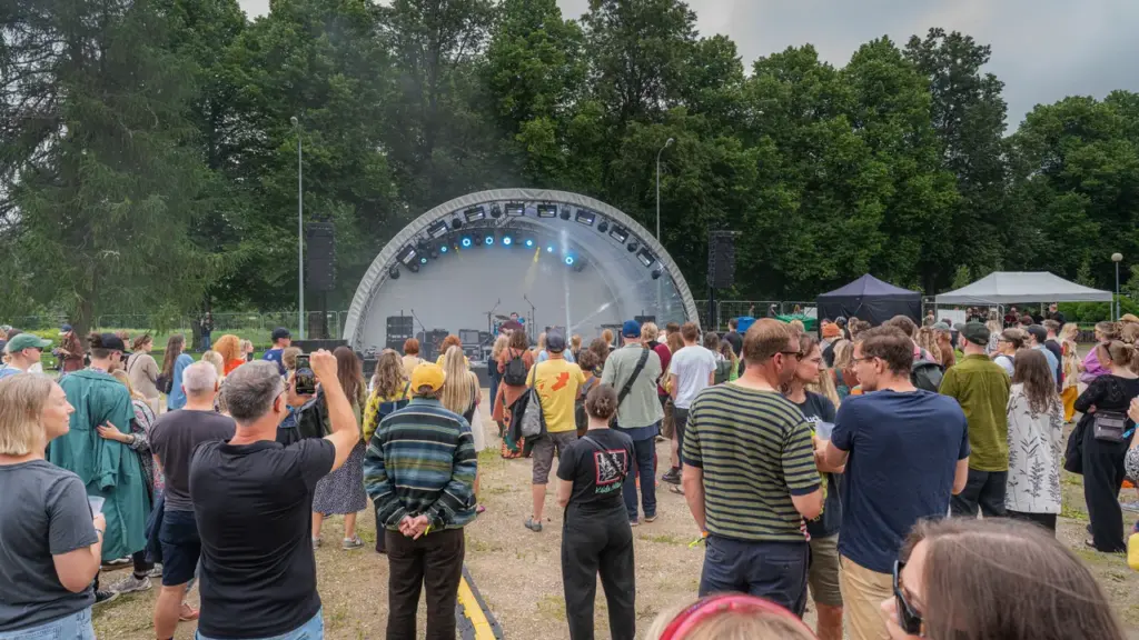 Inimesed vaatavad suvisel vabaõhukontserdil rohelise metsa taustal asuvat kaarekujulist kontserdilava.